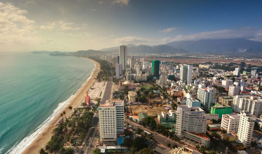 Aerial view of Nha Trang, Vietnam, showing a long sandy beach curving along the turquoise coastline, high-rise buildings, and mountains in the background.