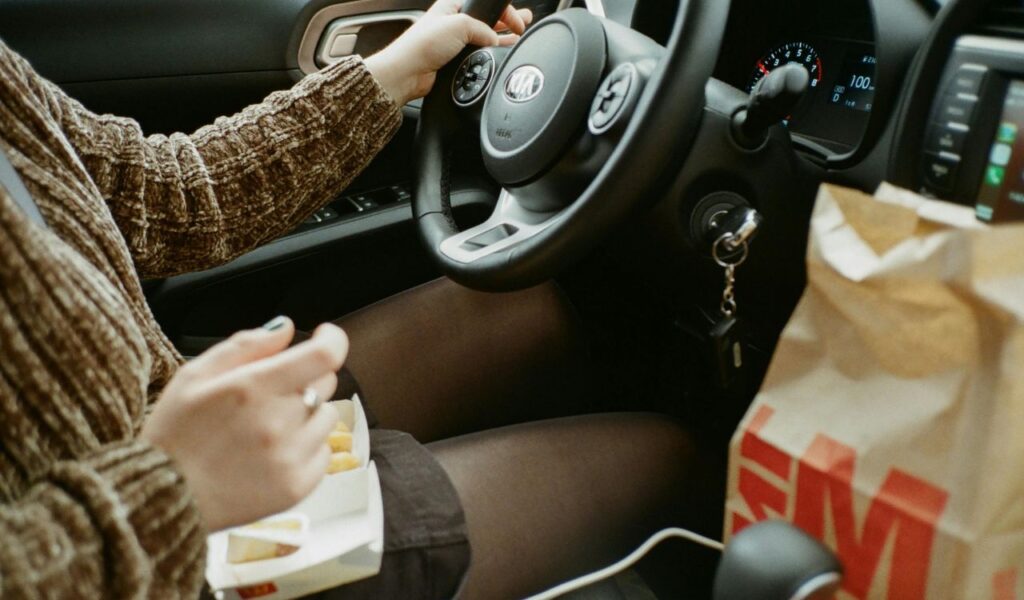 A person driving a car while holding food in one hand, with a bag of fast food on the passenger seat, illustrating eating while driving.