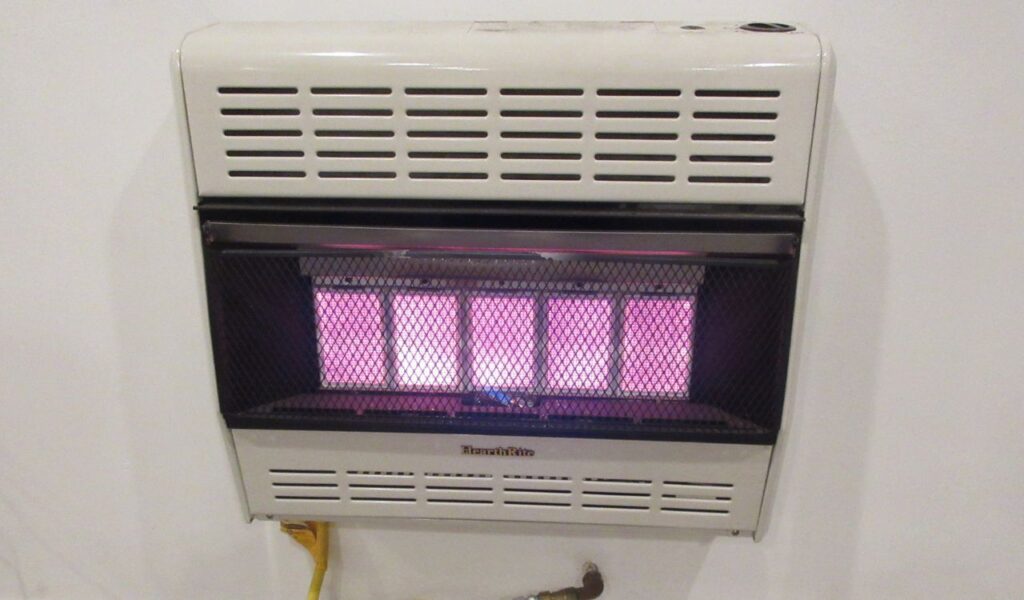A wall-mounted gas heater glows red behind a black mesh screen, providing minimal warmth in a basic indoor setting.
