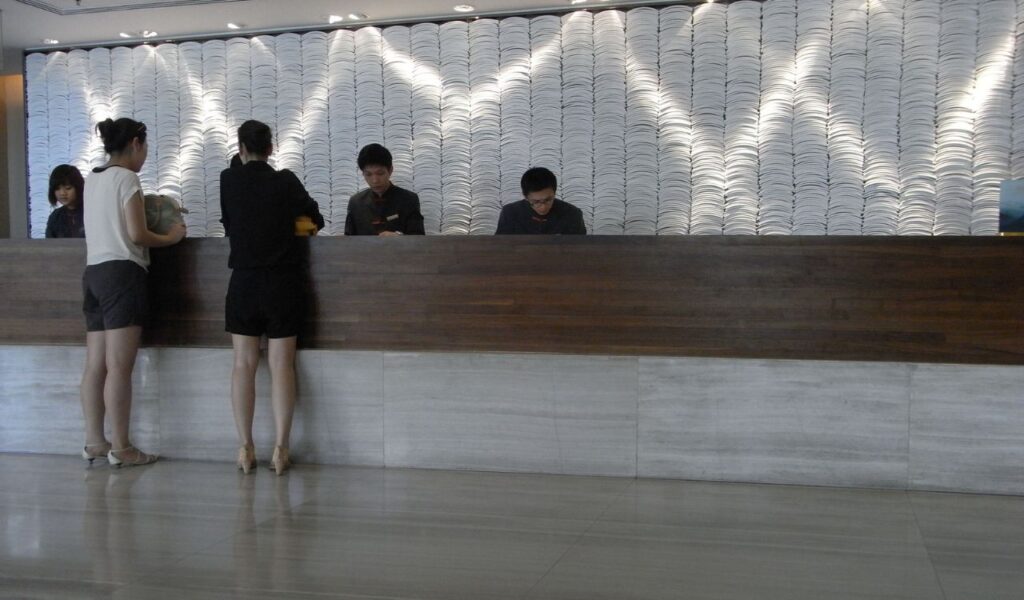 Two guests stand at a modern hotel front desk while three staff members assist with check-in behind a textured white backdrop.