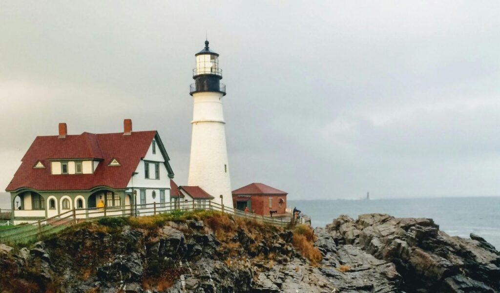 Historic white lighthouse with a red-roofed keeper’s house perched on rocky cliffs, overlooking the ocean under a cloudy sky.
