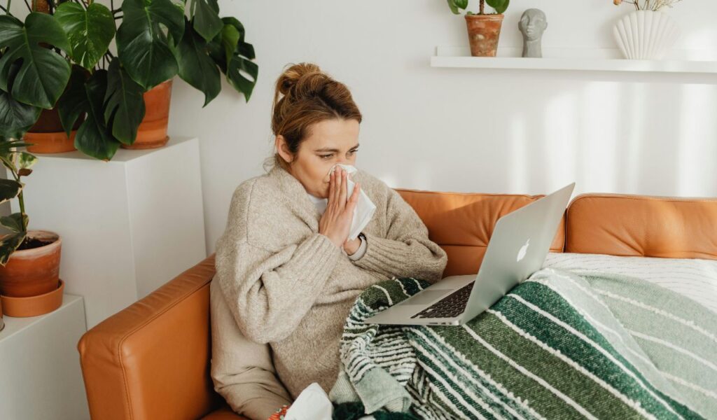 A woman sitting on a couch under a blanket, blowing her nose with a tissue while looking at a laptop, surrounded by potted plants.