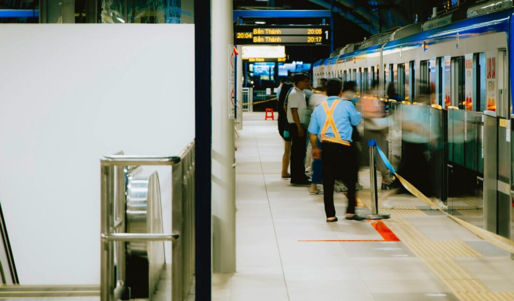 Passengers quickly boarding a train at a station with a platform sign showing departure times, supervised by a staff member in a reflective vest.