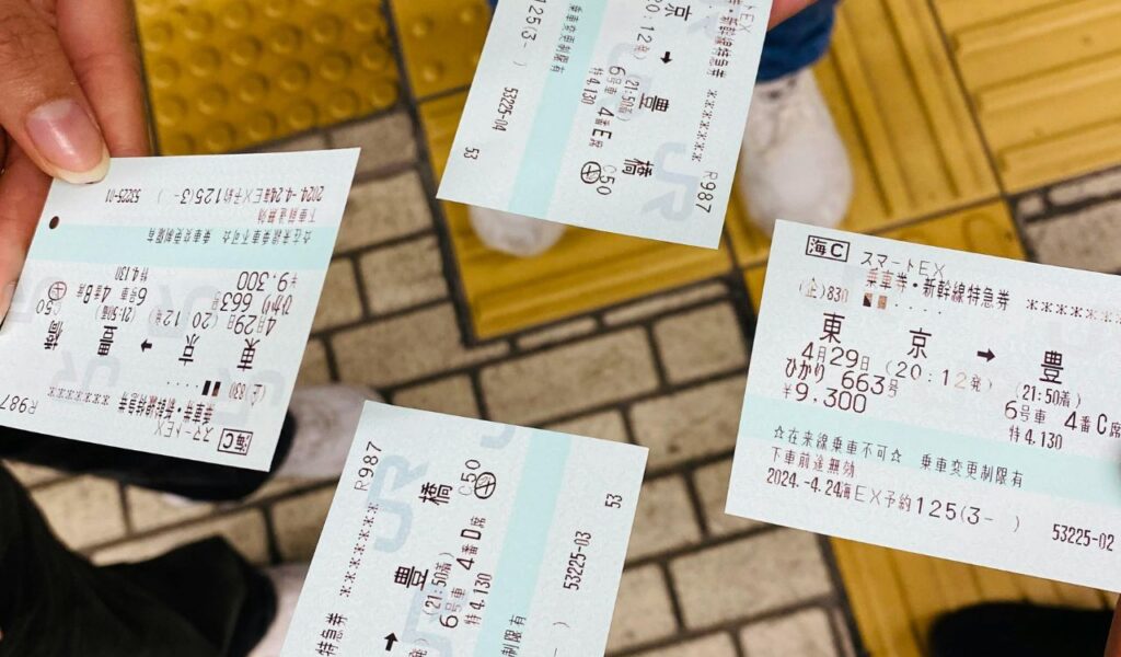 Close-up of four train tickets being held over a tiled platform floor, with travelers' feet visible in the background.