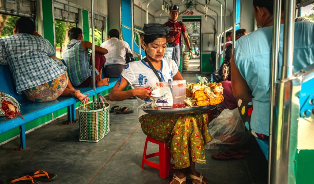 A woman sits on a train seat preparing food with a tray of snacks in front of her, while other passengers sit nearby, creating a lively onboard atmosphere.