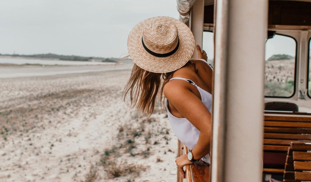 A woman wearing a straw hat leans out of a train window, enjoying a scenic ride along a sandy landscape, capturing the essence of relaxed travel.