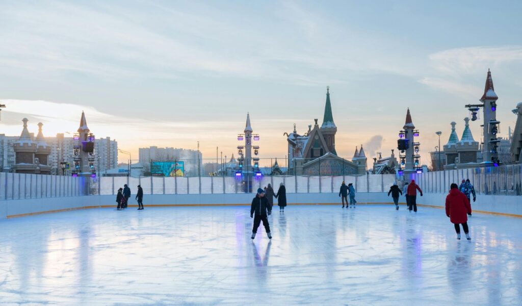 People ice skating at an outdoor rink surrounded by decorative castle-like buildings and colorful lights during winter in the early evening.