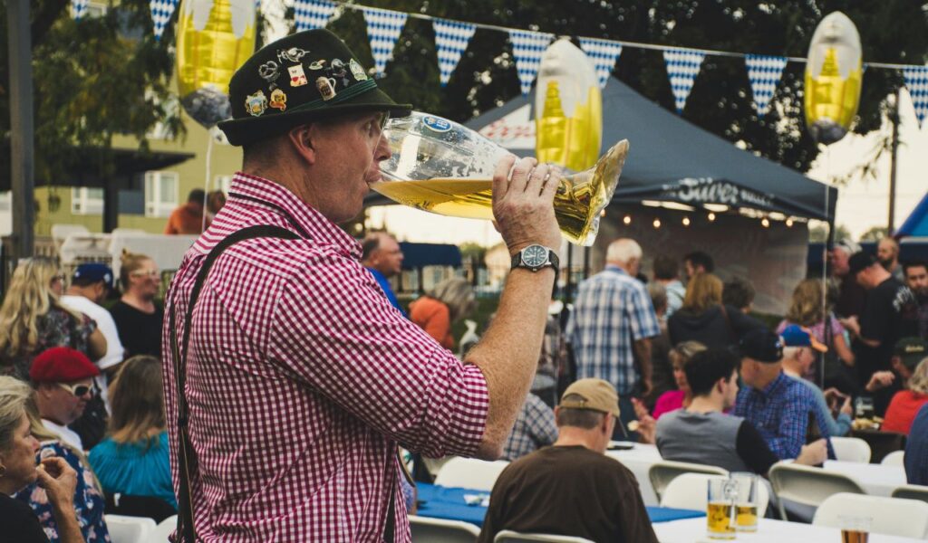 Man in traditional Bavarian attire drinks from a giant beer glass at an Oktoberfest celebration with a lively crowd.