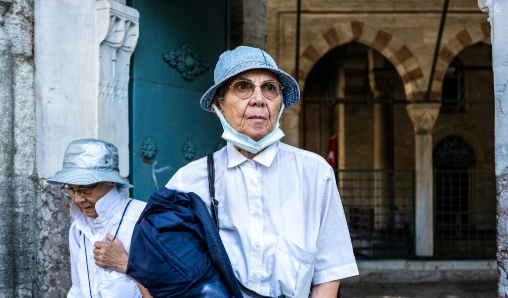 Two older travelers wearing hats and casual clothes walk near a historic building, one carrying a bag and the other adjusting their mask.
