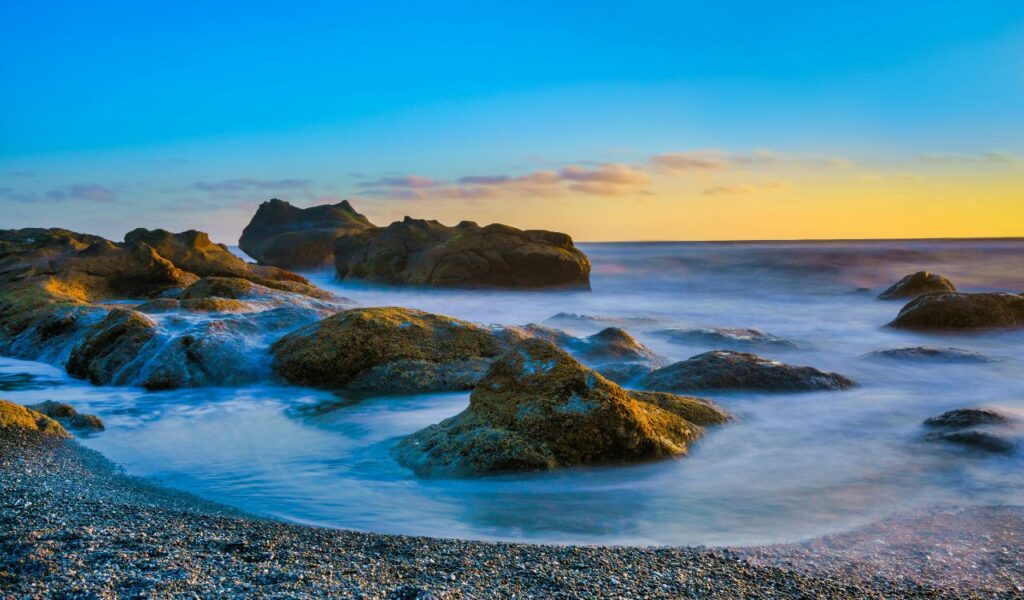 Rocky shoreline at sunset on the Olympic Peninsula, with waves washing over stones and a vibrant blue and orange sky.