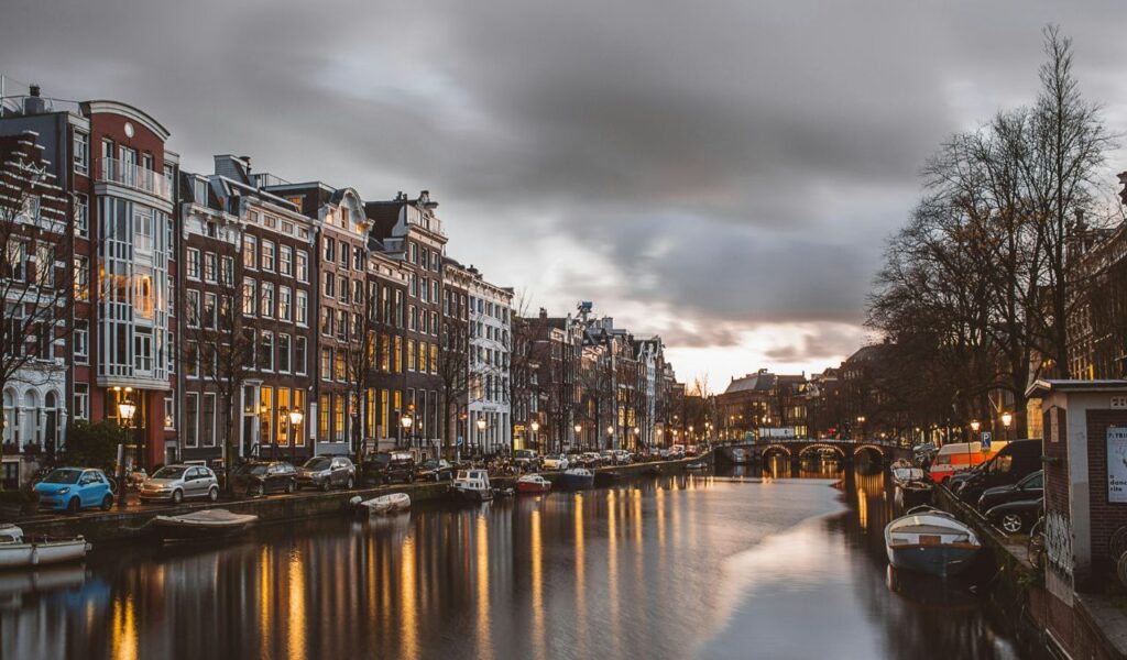 A canal in Amsterdam lined with traditional Dutch buildings, parked cars, and boats, with streetlights reflecting on the water at dusk.