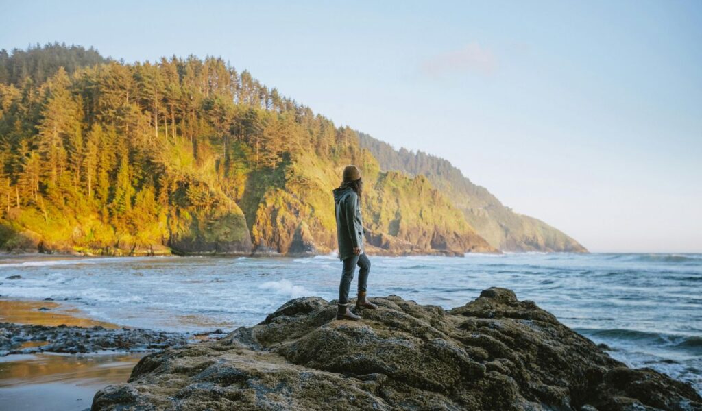 A person in a jacket and beanie stands on a rocky outcrop overlooking the Pacific Ocean, with forested cliffs of the Oregon Coast glowing in golden light.