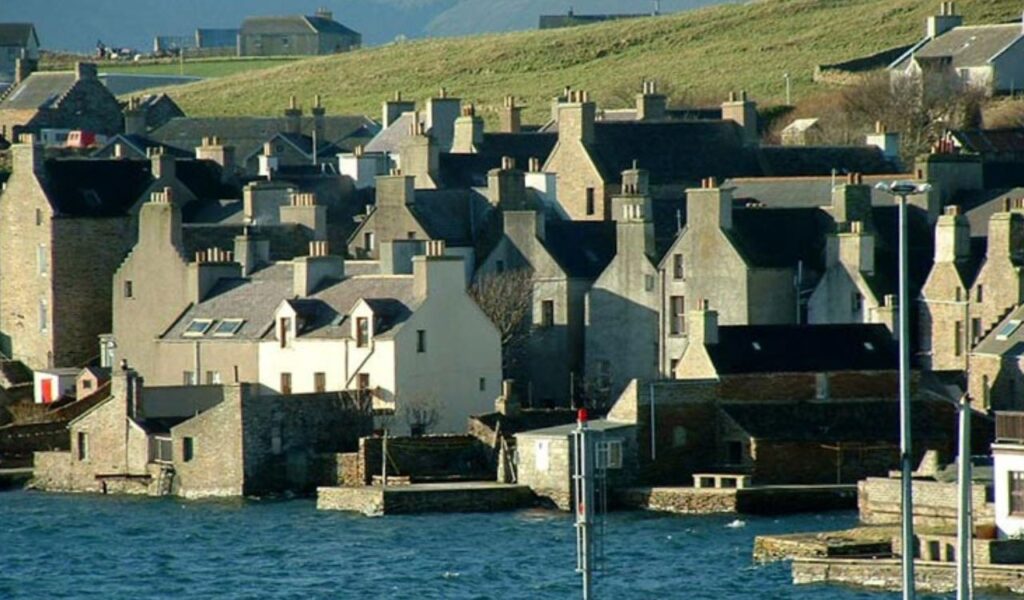 Cluster of old stone buildings with tall chimneys in the Orkney Islands, Scotland, lining a rugged seafront.