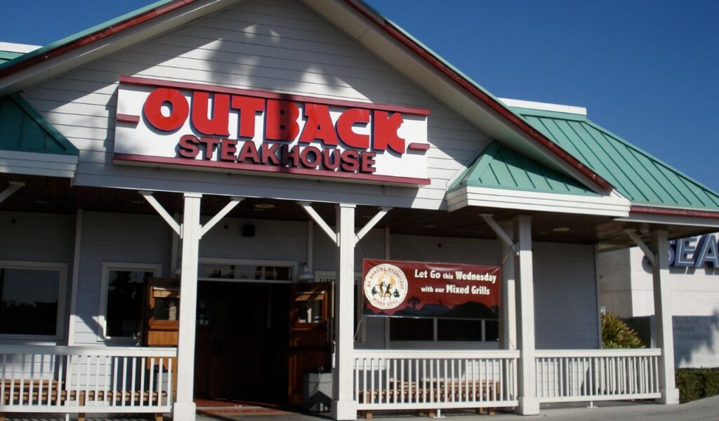 Front view of an Outback Steakhouse with a green roof and promotional banner.