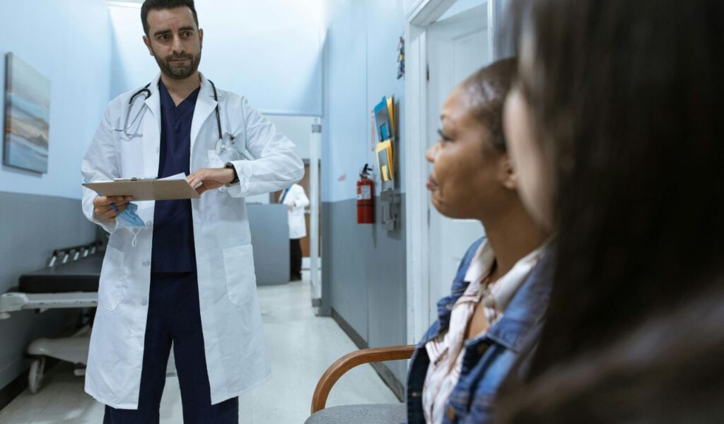 Doctor in white coat reviews a chart while talking with seated patient in a hospital corridor, illustrating overseas medical care.