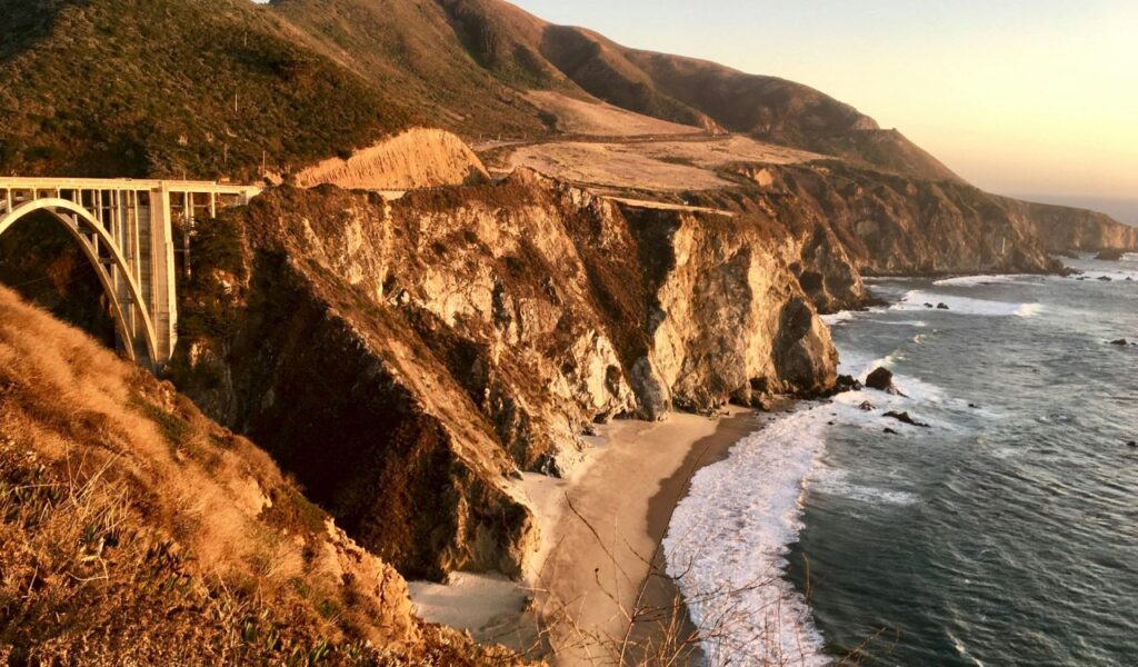Scenic view of the Pacific Coast Highway with a bridge spanning rugged cliffs, overlooking the ocean waves crashing onto a sandy beach below.