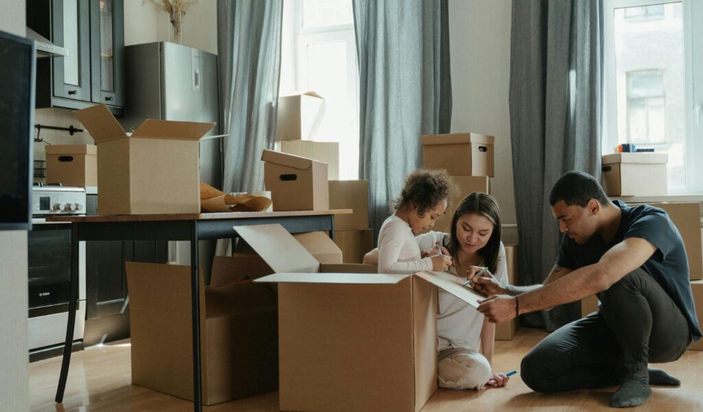 A family sits on the floor of a home surrounded by cardboard boxes, labeling and organizing items during a move or packing session.