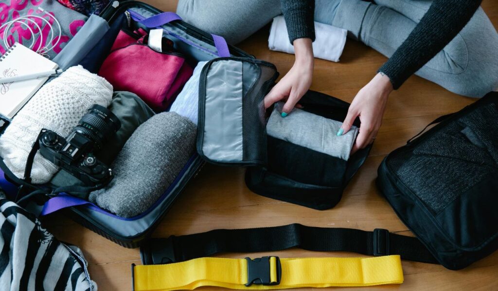 A person organizing clothes, camera, and travel essentials into a suitcase, preparing for a trip with packing cubes and accessories.