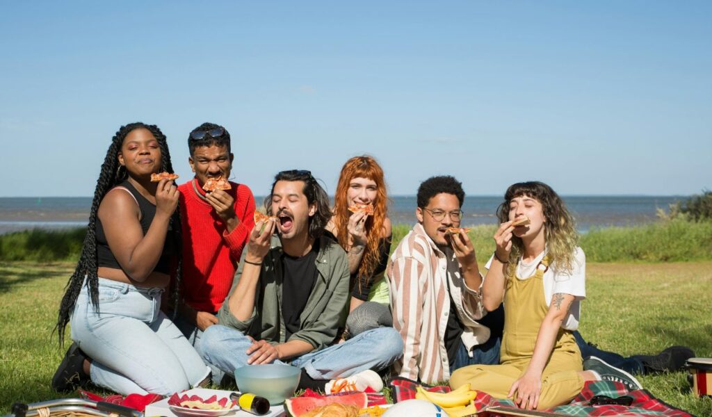 A group of friends enjoys a picnic on a grassy field near the water, sitting on a blanket and happily eating slices of pizza under a clear blue sky.