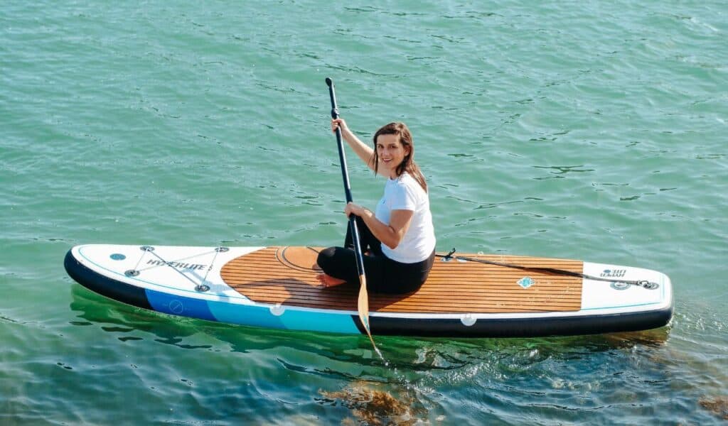 Woman sitting on a stand-up paddleboard with a paddle in hand, smiling while floating on calm, clear green water.