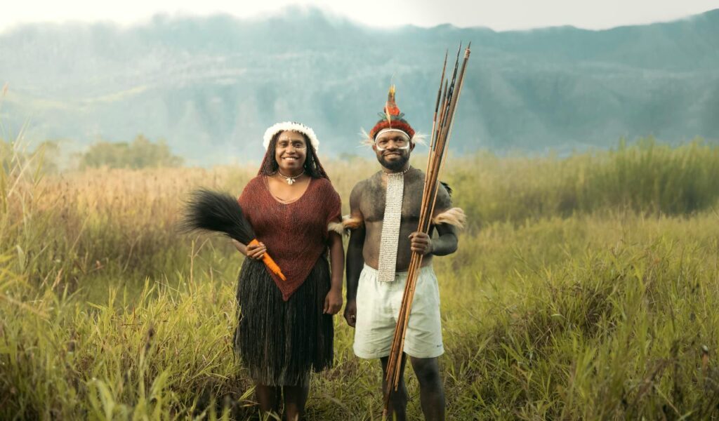 A man and woman in traditional Papua New Guinea attire stand smiling in a grassy field with mountains behind them.