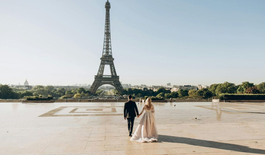 A couple holds hands while walking toward the Eiffel Tower in Paris on a clear day, with a scenic city view in the background.
