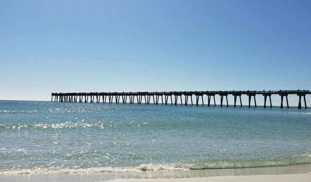 Pensacola Beach pier extending over calm turquoise waters with gentle waves lapping the sandy shore under a clear blue sky.