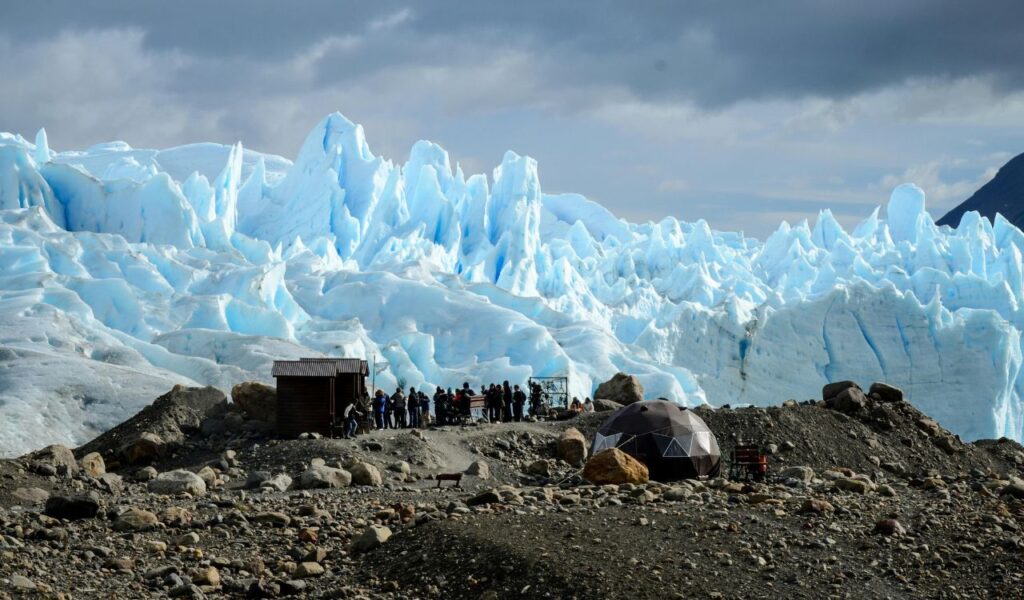 Perito Moreno Glacier in Argentina with towering blue ice formations and visitors gathered on rocky terrain in the foreground.