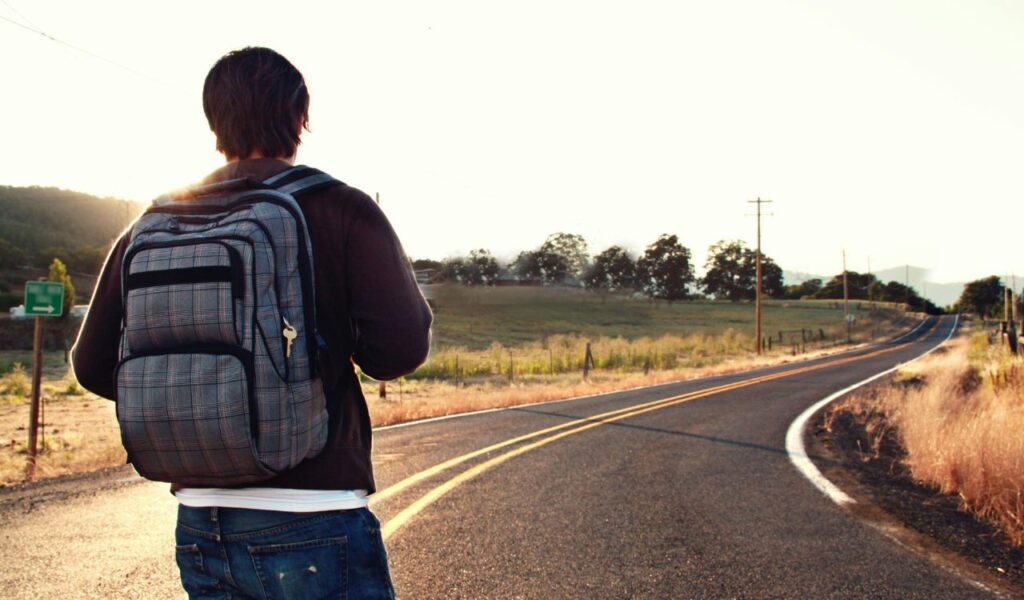 A solo traveler with a large backpack stands on a rural road at sunrise, facing a winding path through open countryside.