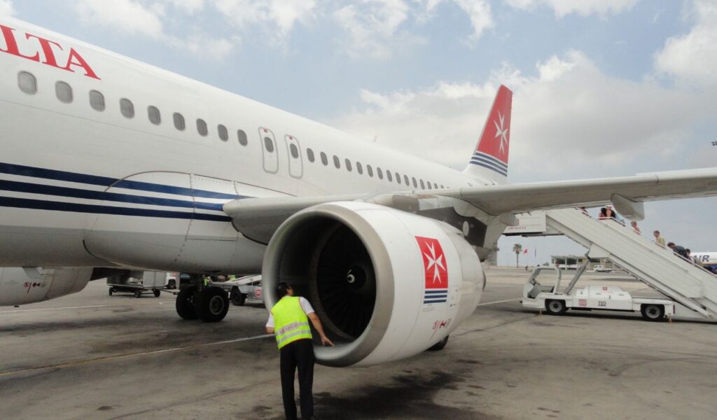 Ground crew member inspects an Air Malta jet engine on the tarmac, with passengers boarding via mobile stairs nearby.