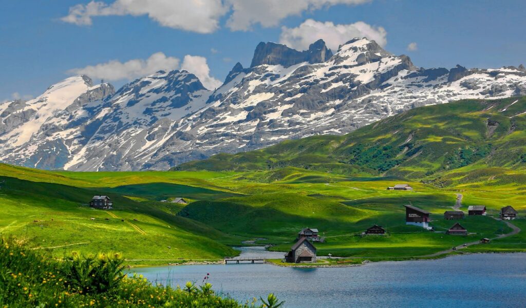 Scenic view of Swiss Alps with snow-covered peaks, green rolling hills, alpine cottages, and a calm blue lake in the foreground.