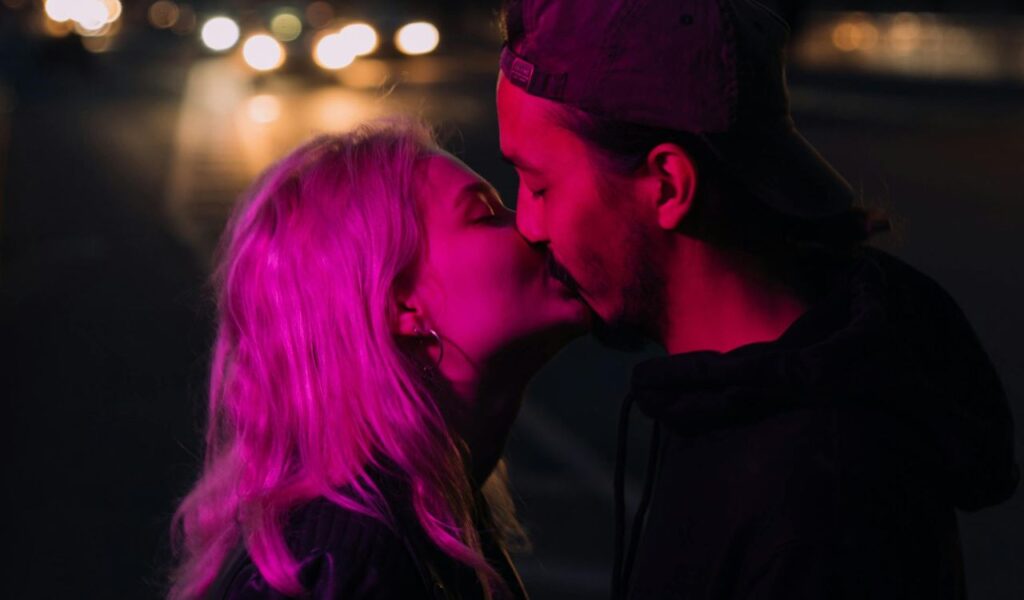 A couple sharing a kiss under pink neon lighting at night with blurred city lights in the background.