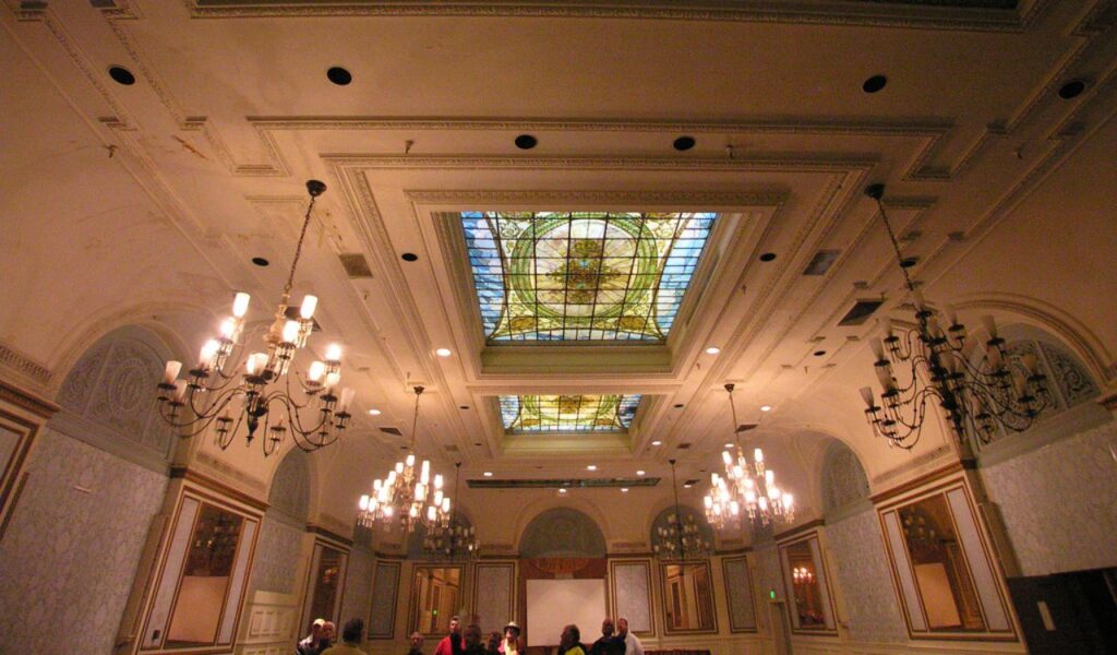 Guests gather and chat beneath ornate chandeliers and stained glass skylights in a grand, echoing hotel ballroom.