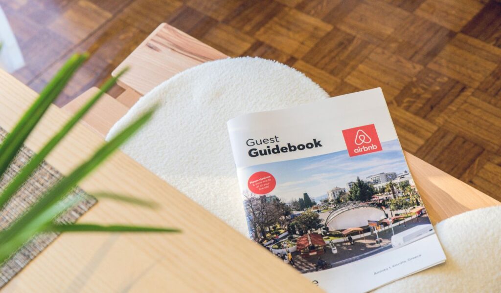 A printed Airbnb guest guidebook rests on a light wooden table, showcasing travel tips and a cover photo of a sunny cityscape.