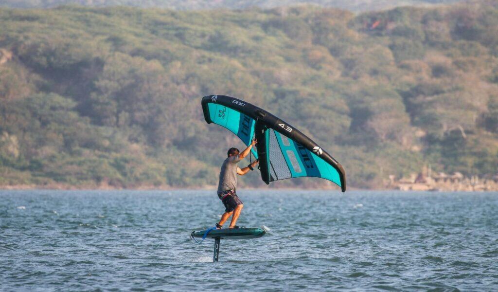 A windsurfer gliding over Floras Lake on a foil board with forested hills in the background