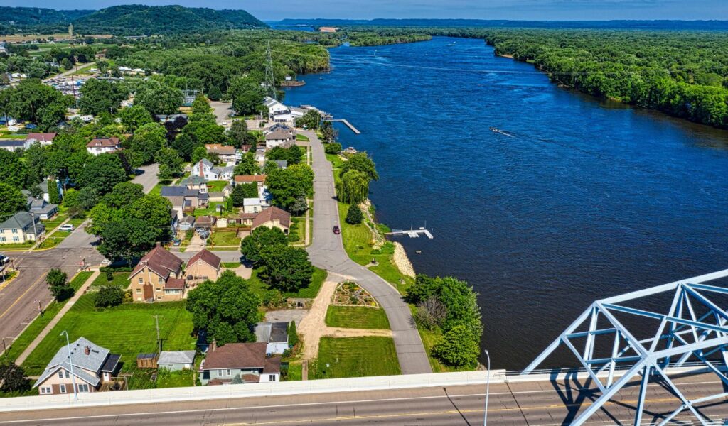 Aerial view of a small riverside town with neat homes, green trees, and a winding road leading to a large blue river, with a steel bridge in the foreground and forested hills in the distance.