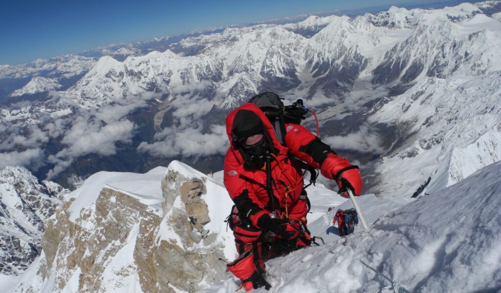 A climber in red cold-weather gear ascends a steep snow-covered mountain ridge, surrounded by a vast range of rugged, snow-capped peaks.