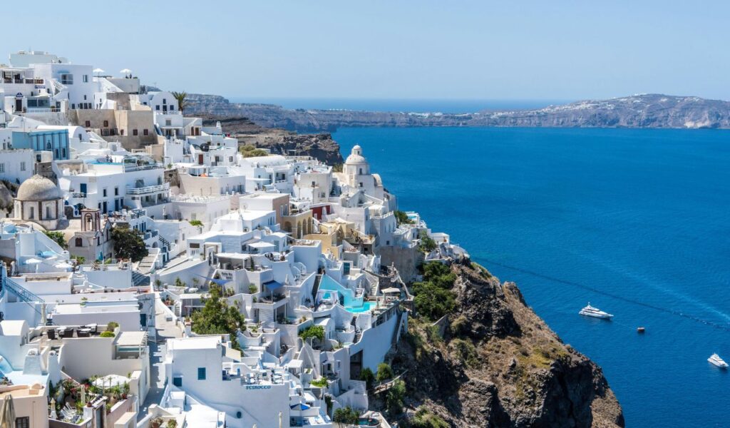 A cliffside view of whitewashed buildings in Santorini overlooking the deep blue Aegean Sea, with boats scattered across the water under a clear sky.