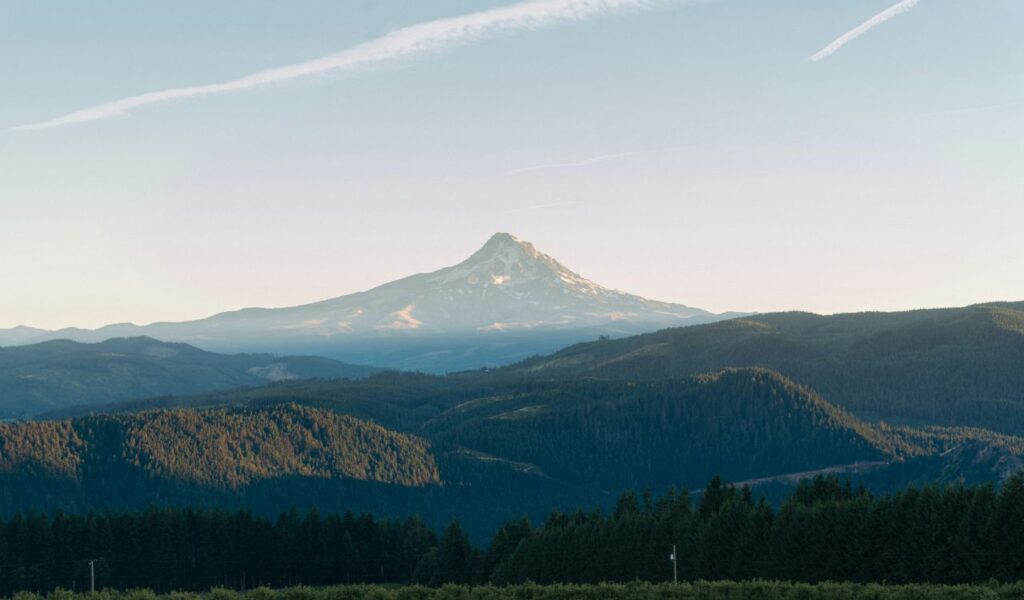  A majestic peak rising above layers of forest-covered hills, viewed under a clear, pale sky at dusk.