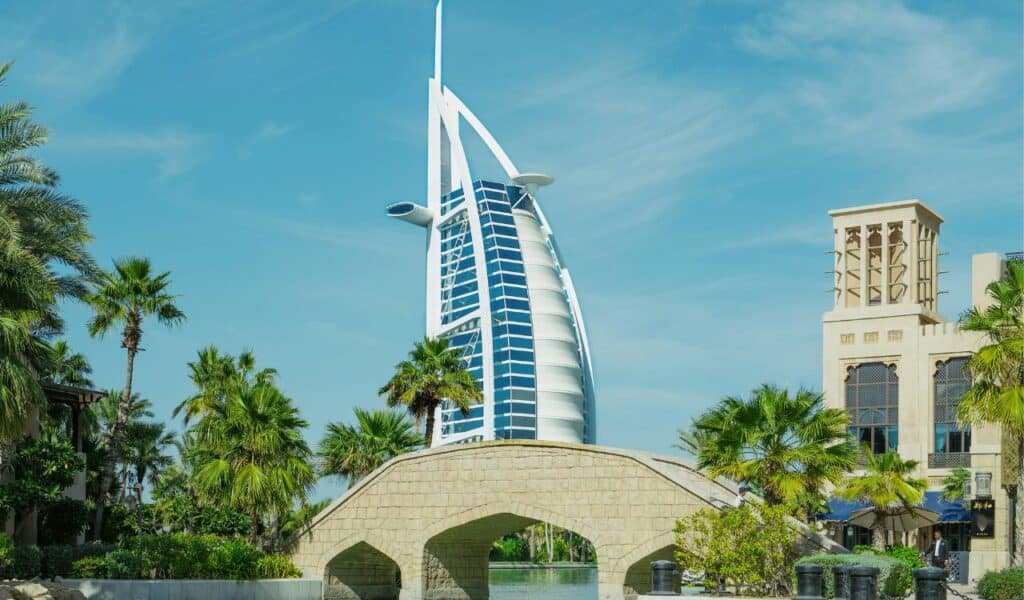 Daytime view of the iconic Burj Al Arab hotel in Dubai, shaped like a sail, surrounded by palm trees and traditional Arabian architecture, with a stone bridge in the foreground.