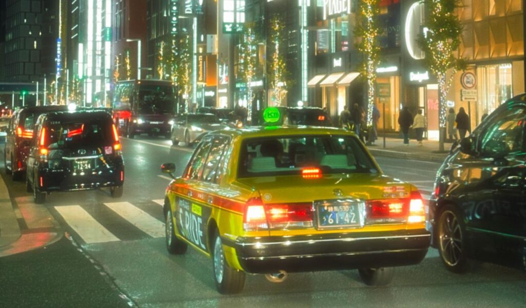Cars driving along a brightly lit city street at night, with illuminated buildings, shops, and festive lights lining the road.