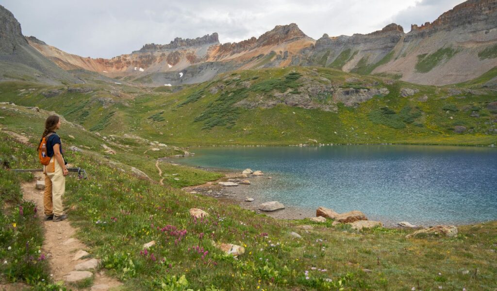 A hiker overlooks a serene alpine lake surrounded by lush meadows and rugged peaks along the San Juan Skyway in Colorado.