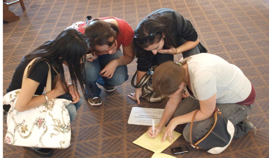 Four women kneel on a carpeted floor, focused on solving a scavenger hunt clue written on yellow and white paper.