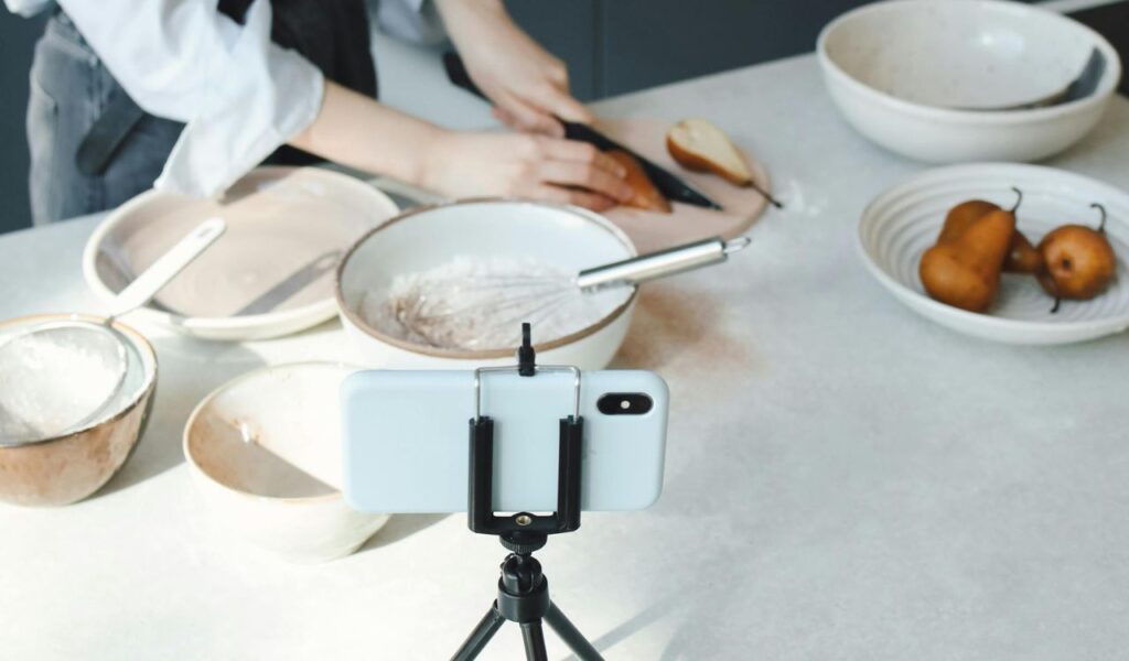A person chopping a pear on a cutting board while filming a cooking tutorial with a smartphone on a tripod, surrounded by bowls and baking ingredients.