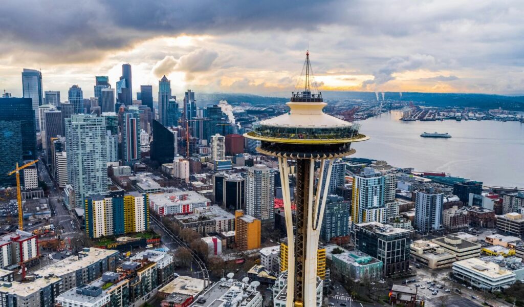 Aerial view of Seattle, Washington featuring the iconic Space Needle in the foreground, with the city skyline, Puget Sound, and cloudy sky in the background.
