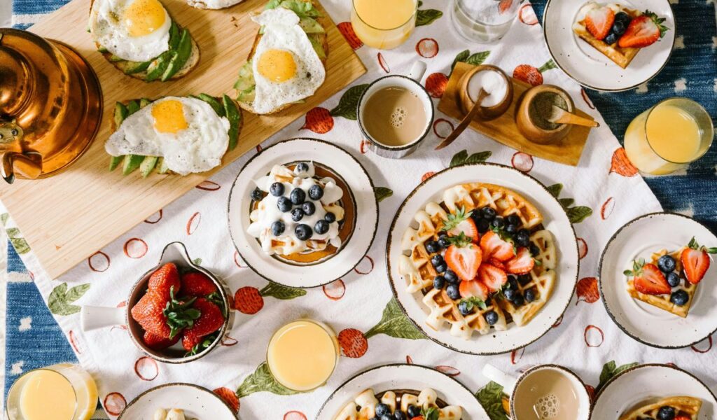 A vibrant breakfast spread featuring waffles topped with fresh strawberries and blueberries, avocado toast with eggs, coffee, and orange juice on a fruit-patterned tablecloth.