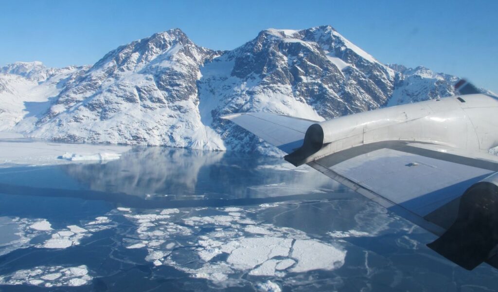 Aerial view of Sermilik Fjord in Greenland, with icy water, floating ice chunks, and snow-covered mountains below.