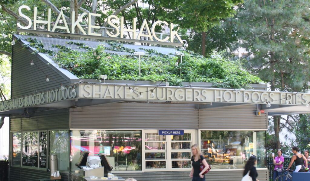 Outdoor Shake Shack location surrounded by greenery, featuring a walk-up window and a “Pick Up Here” sign, with people passing by.