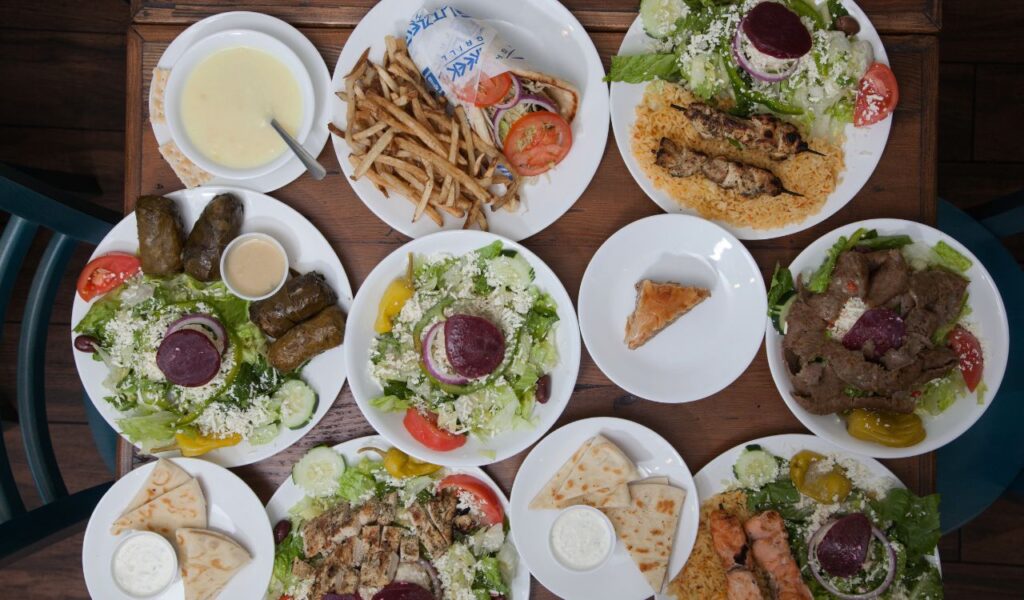 Overhead view of a wooden table filled with assorted Greek dishes, salads, pita, grape leaves, fries, and grilled meats meant for sharing.