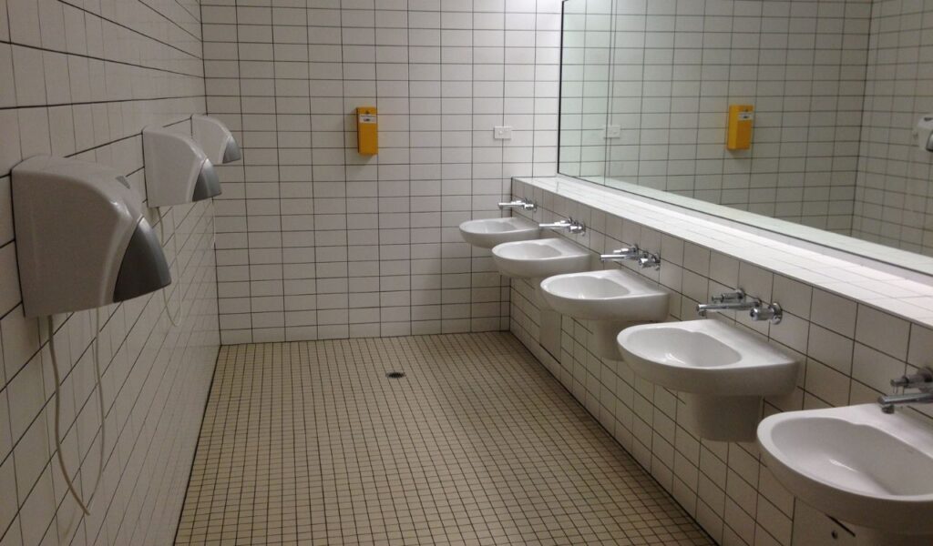 A tiled communal bathroom features a long wall of sinks with mirrors and hand dryers, evoking a basic shared guesthouse setup.
